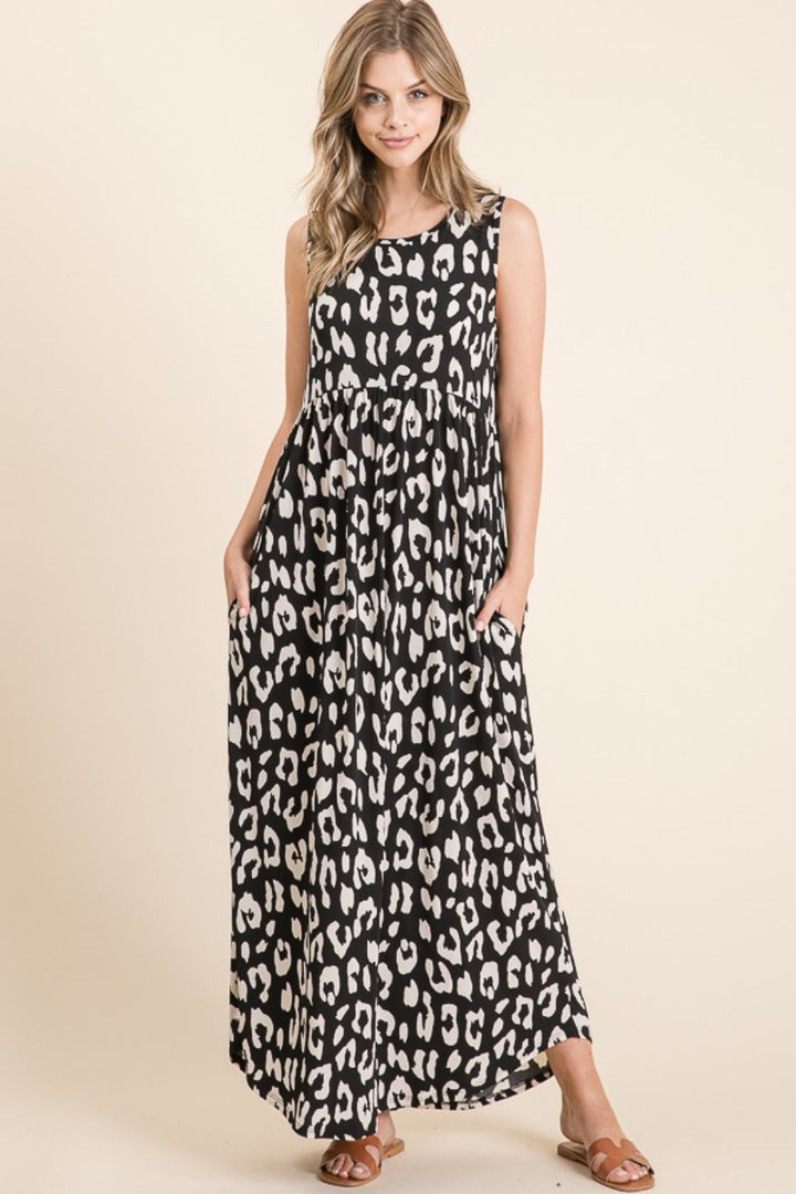 A woman stands against a beige background in a BOMBOM Leopard Maxi Dress with Pockets, featuring a sleeveless black and white leopard print and a shirring waist. Her long hair cascades as she smiles softly with hands in pockets, completing her stylish look with brown sandals.