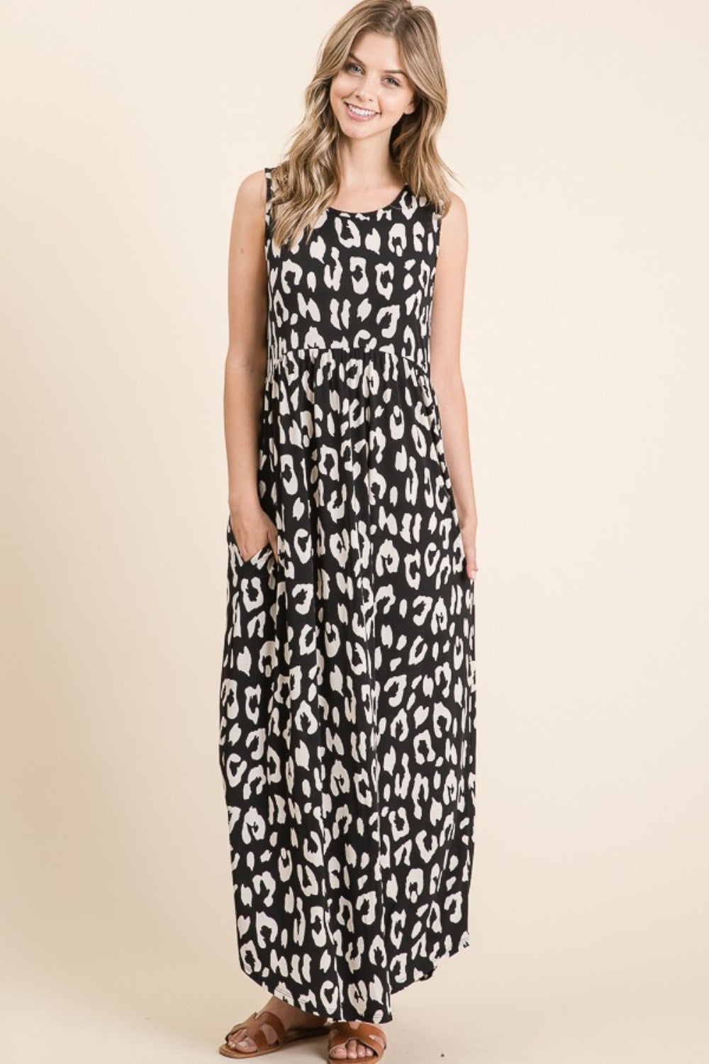 Smiling, a person stands in a BOMBOM Leopard Maxi Dress with pockets, the sleeveless shirring waist design cascading elegantly, wearing brown sandals against a neutral background.