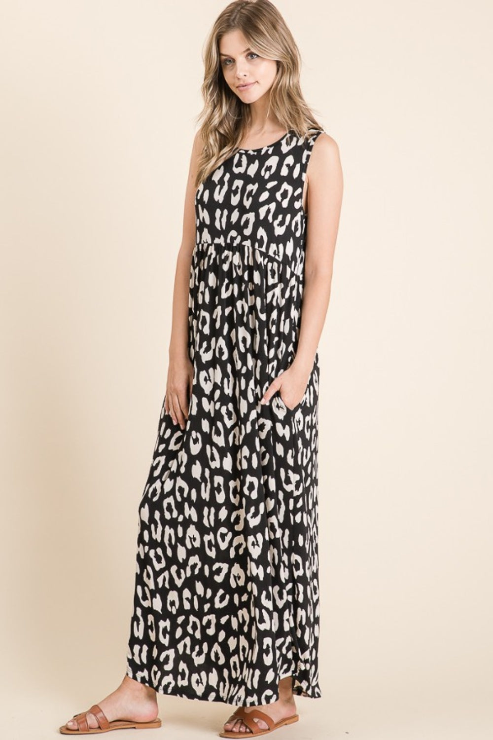 A woman in a BOMBOM sleeveless black and white leopard maxi dress with pockets stands against a beige background. She has long hair, wears brown sandals, and looks toward the camera.