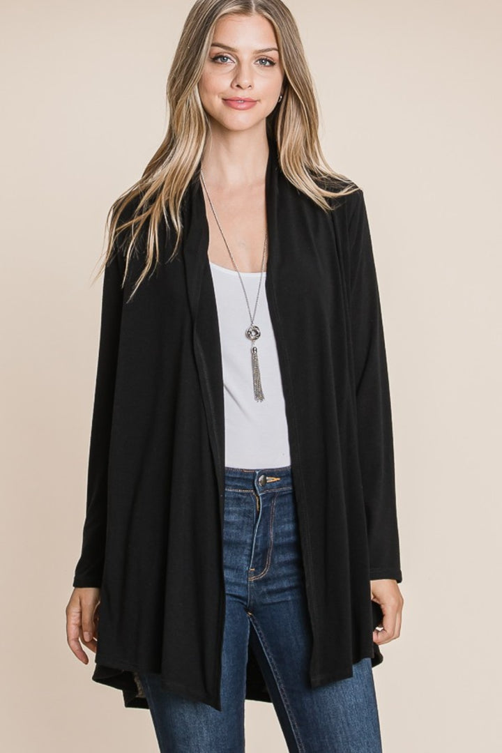 A person with long hair is wearing the BOMBOM Open Front Long Sleeve Cardigan in black over a white top and blue jeans, accessorized with a long pendant necklace, standing against a plain beige background.