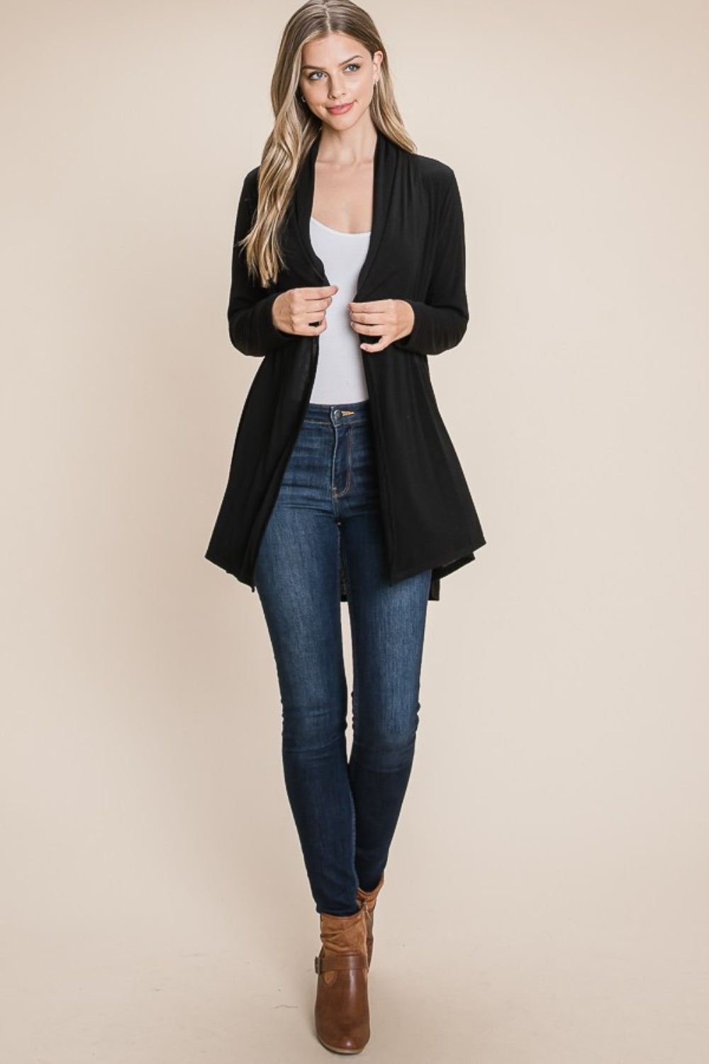 A person with long hair wears the chic BOMBOM Open Front Long Sleeve Cardigan in black over a white top, blue jeans, and brown ankle boots. They stand against a beige background, smiling softly with hands gently gripping the cardigan.