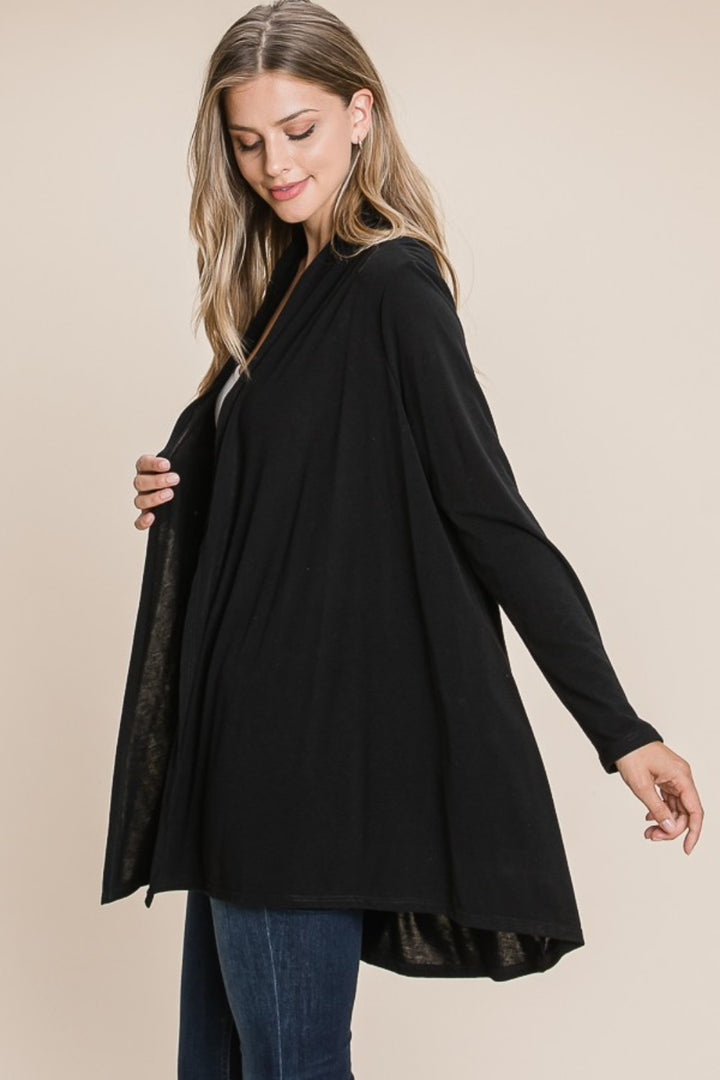 A woman with long hair is wearing a BOMBOM Open Front Long Sleeve Cardigan, an ideal layering piece made of soft knit. She stands against a neutral background, gently holding one side of the cardigan while looking down with a gentle smile.