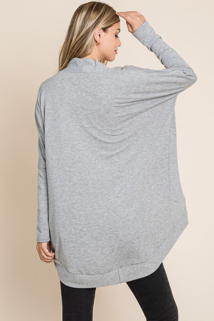 A woman in a versatile BOMBOM Open Front Long Sleeve Cocoon Cardigan paired with black pants stands facing away. Her hand touches her head, revealing glimpses of blonde hair. The plain beige background complements the cozy elegance of her ensemble.