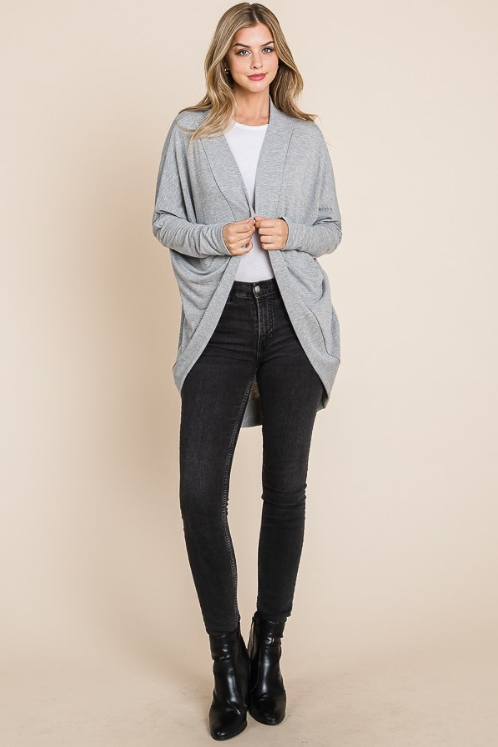 A person stands against a neutral background wearing a BOMBOM Open Front Long Sleeve Cocoon Cardigan in gray, paired with a white top, black jeans, and black ankle boots. Their hands gently hold the edges of this versatile layering piece.