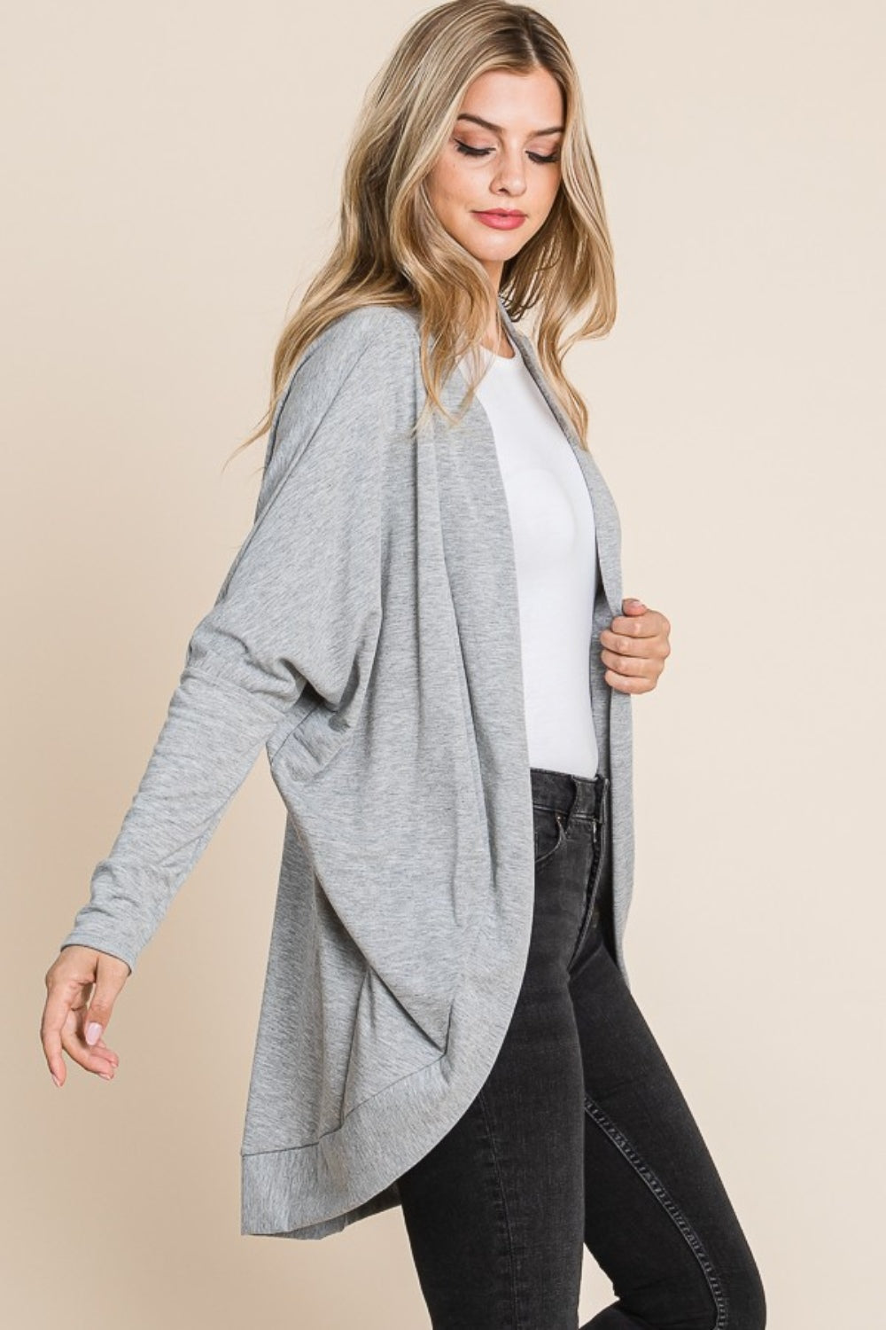 A person with long hair wears a BOMBOM Open Front Long Sleeve Cocoon Cardigan in light gray over a white top and black pants. This versatile piece complements the plain, light beige background as they stand and look down slightly to the side.