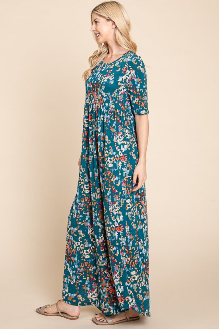 A woman with long blonde hair wears the BOMBOM Printed Shirred Maxi Dress, featuring a colorful floral pattern on a teal background. She stands sideways against a beige backdrop in sandals, smiling and showcasing this versatile statement piece.