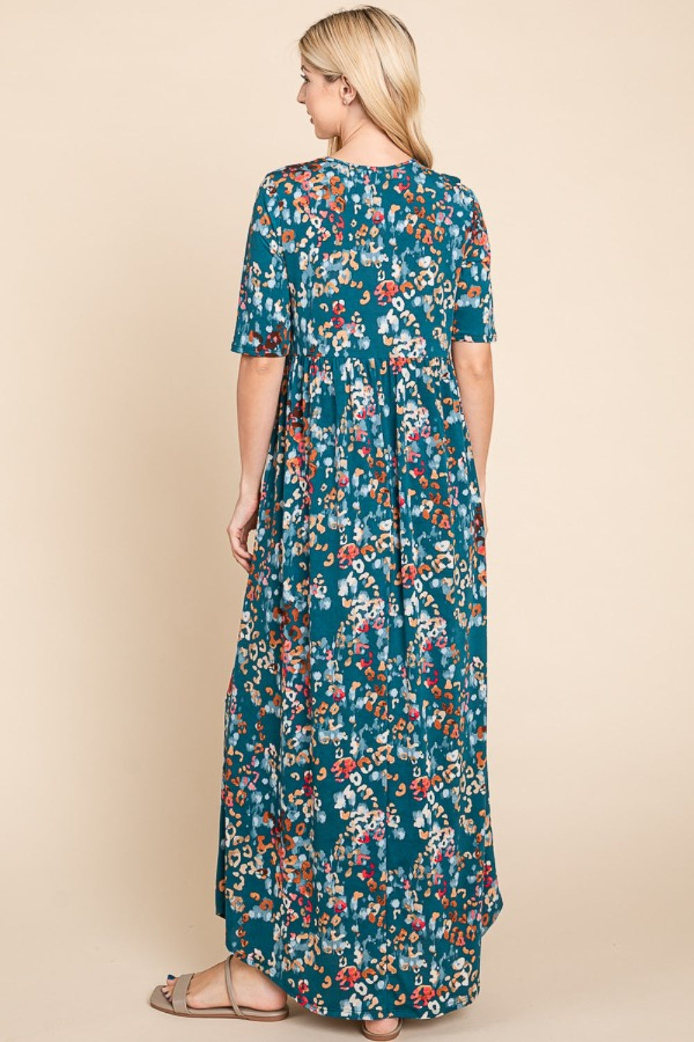 A blonde woman models the BOMBOM Printed Shirred Maxi Dress, a floral-patterned blue garment. She faces away, displaying its flowing elegance against a plain background.