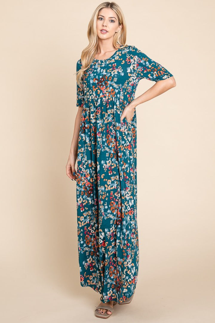 Against a beige backdrop, a woman poses confidently in a BOMBOM Printed Shirred Maxi Dress adorned with red, white, and yellow flowers on teal fabric. Her long blonde hair enhances the dress as she stands with one hand on her hip and the other by her side.