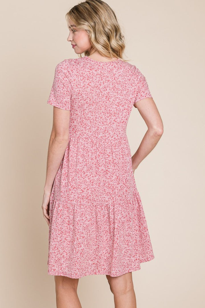 A woman with wavy blonde hair is seen from the back wearing a BOMBOM Printed Short Sleeve Mini Dress. The pink floral summer dress features a relaxed fit and a tiered skirt, set against a neutral beige background that enhances the serene atmosphere.