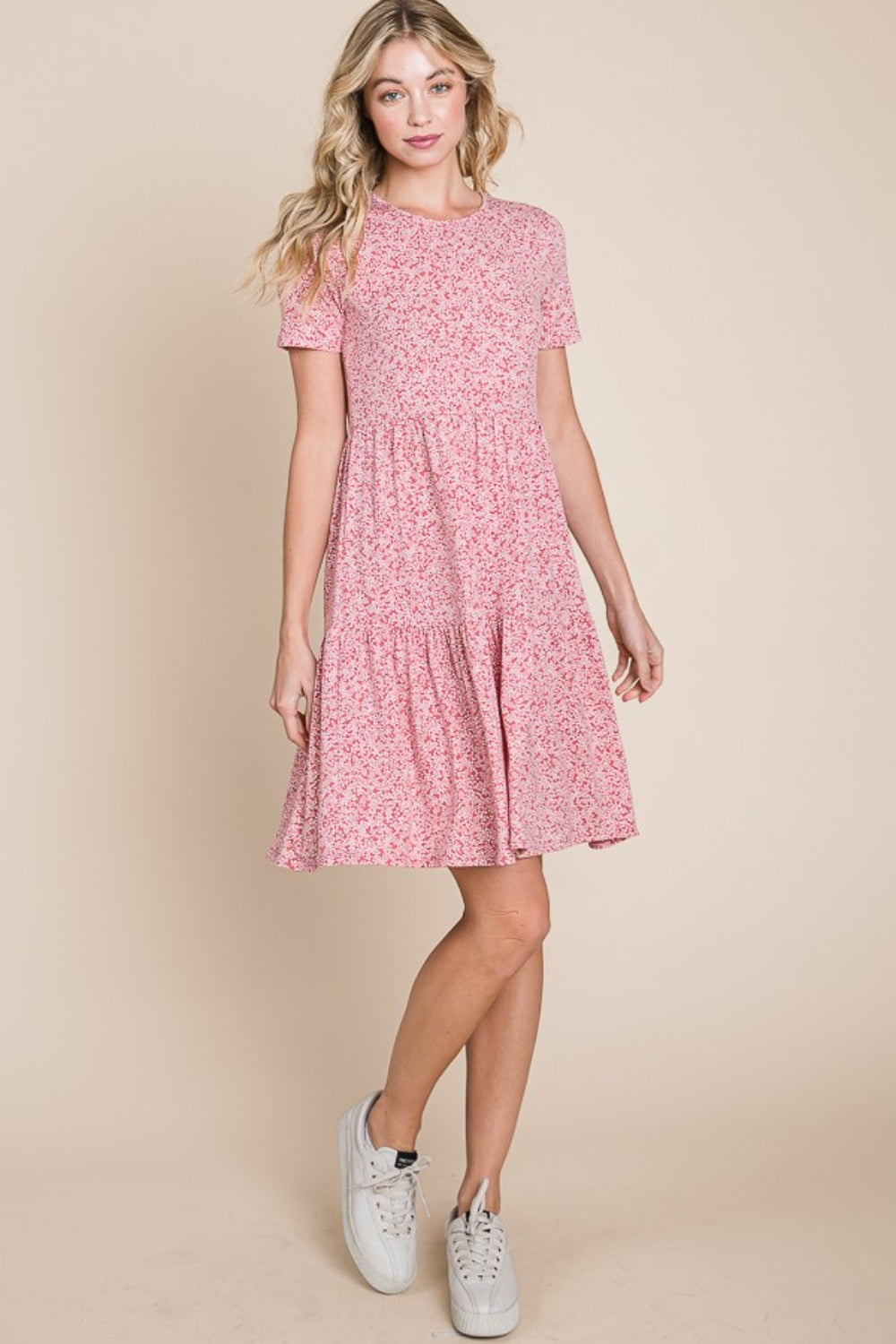 Someone in a pink BOMBOM Printed Short Sleeve Mini Dress and white sneakers stands against a beige background. They have long blonde hair, a neutral expression, one hand resting beside them, and the other slightly raised while looking at the camera.