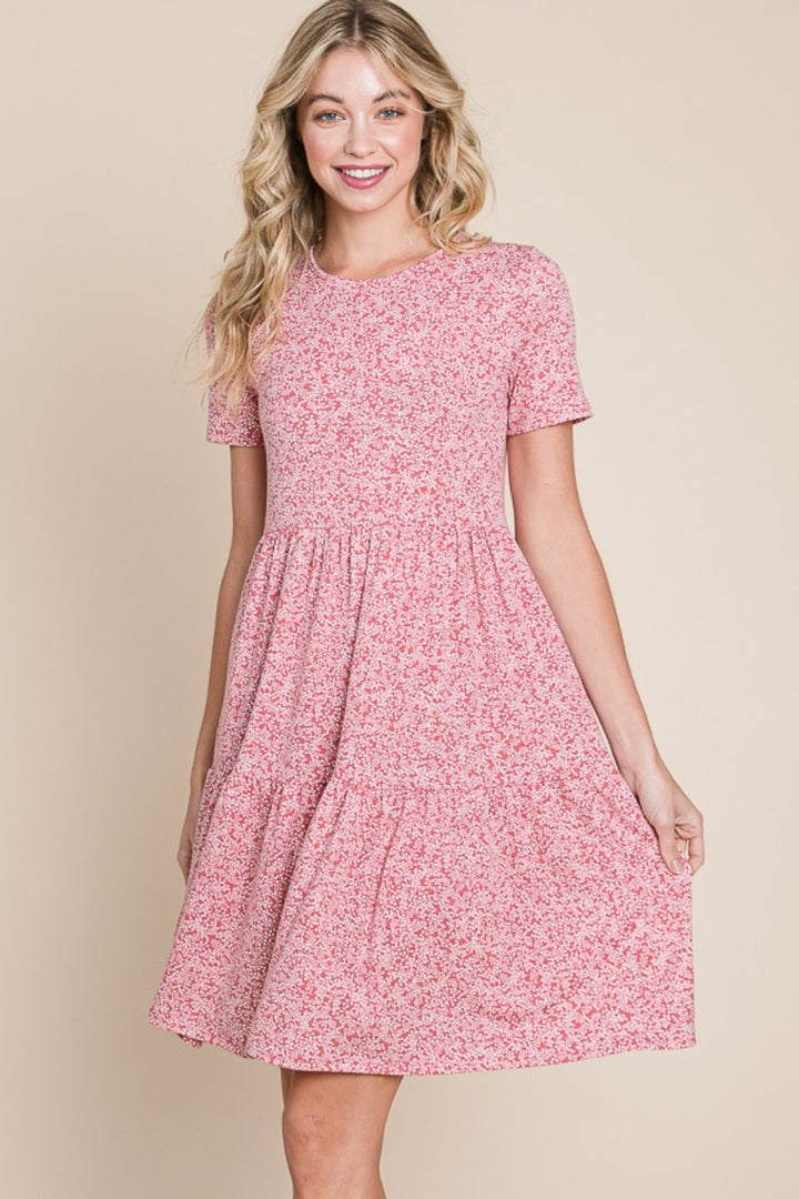 A person smiles in front of a beige backdrop, wearing the BOMBOM Printed Short Sleeve Mini Dress with a pink floral design, featuring a gathered waist and flowing skirt—ideal as a lightweight summer dress.