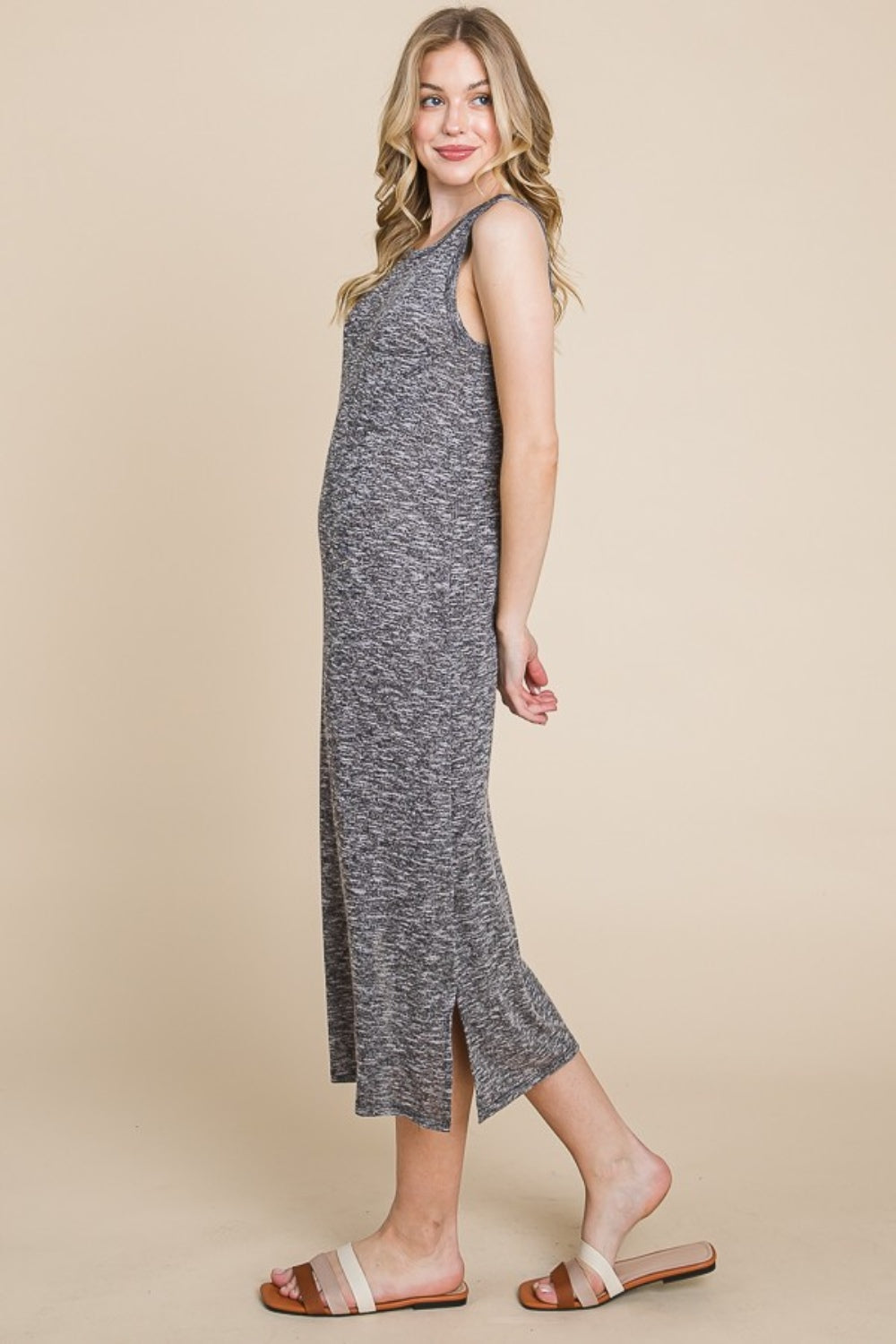 Against a neutral backdrop, a person with long hair stands sideways, exuding chic elegance in a BOMBOM Round Neck Slit Midi Tank Dress and white sandals. Their slight smile enhances this perfect summer wardrobe addition.