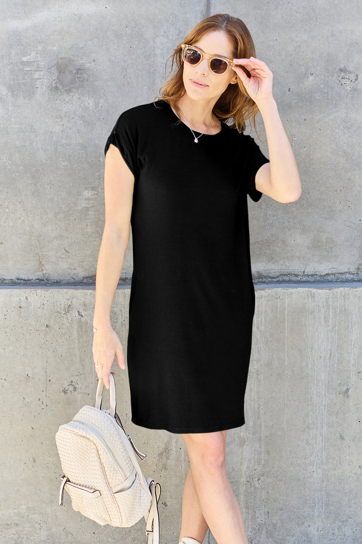 A woman in a black Basic Bae Bamboo dress poses confidently against a concrete backdrop, wearing sunglasses. She holds a textured white bamboo backpack and smiles, brushing her hair back with one hand.