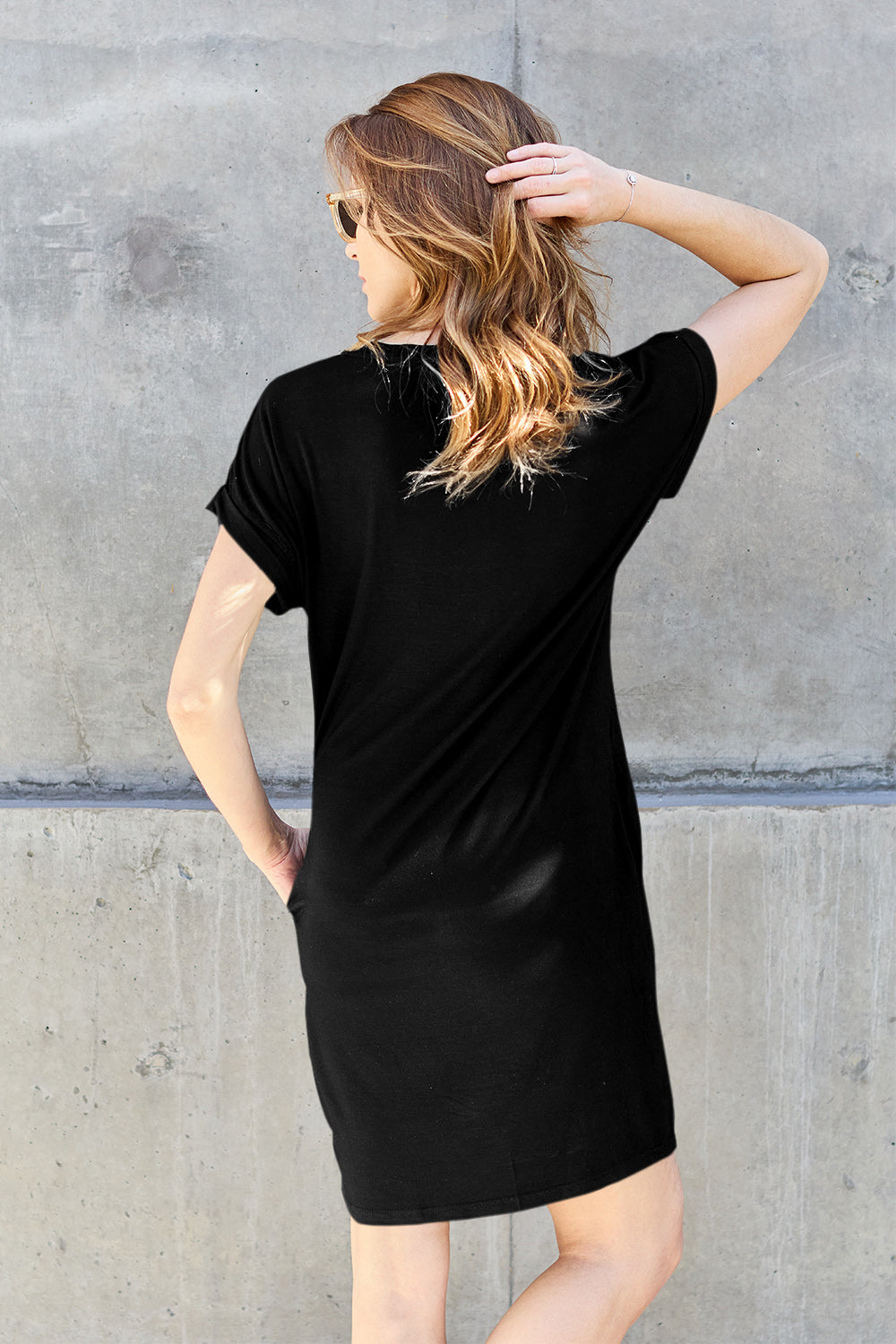 A woman with wavy blond hair stands against a concrete wall, wearing the Basic Bae Bamboo Full Size Round Neck Short Sleeve Dress in black. She touches her hair with one hand, while the other is in the dresss pocket, showcasing its modern, machine wash cold design.