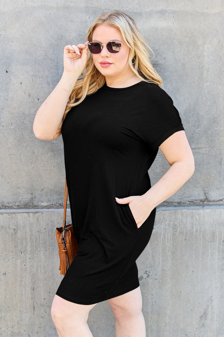 A person with long blonde hair stands against a gray concrete wall wearing sunglasses and the Basic Bae Bamboo Full Size Round Neck Short Sleeve Dress with pockets. They hold a small brown purse, one hand in a pocket, while the other adjusts their sunglasses.