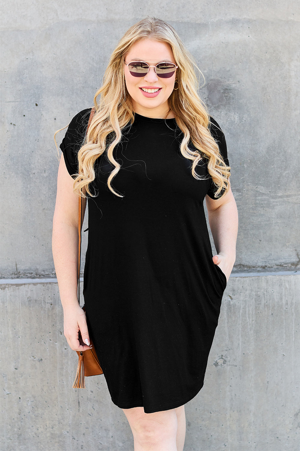 A person with long, wavy blonde hair wears sunglasses and the Basic Bae Bamboo Full Size Round Neck Short Sleeve Dress with Pockets in black. They smile in front of a concrete wall, hand in pocket and a tan handbag over their shoulder.