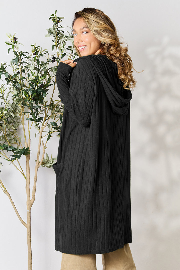 Wearing the Basic Bae Full Size Hooded Sweater Cardigan, a person with long hair smiles while turning toward the camera. The black, ribbed cardigan is paired with beige pants and boasts an opaque stretch design that is ideal for machine wash cold. They are standing beside a small leafy plant against a light, neutral background.