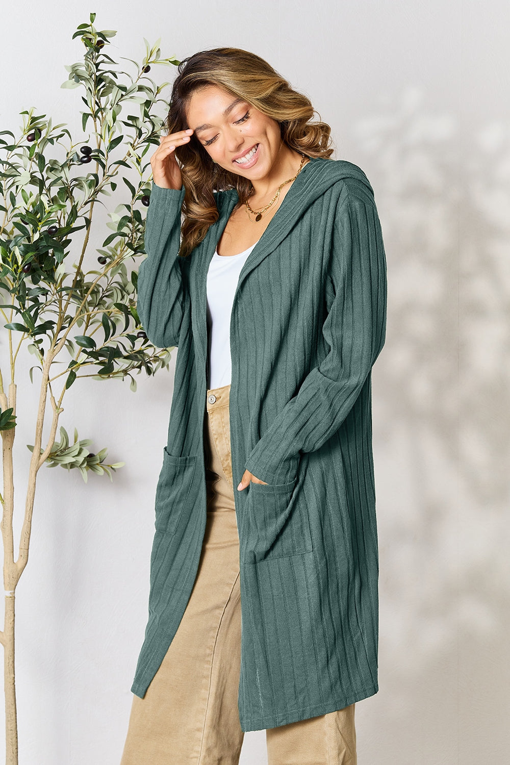 A person smiling and touching their head is wearing the Basic Bae Full Size Hooded Sweater Cardigan in teal, layered over a white top and beige pants. They stand beside a small leafy plant against a light background.