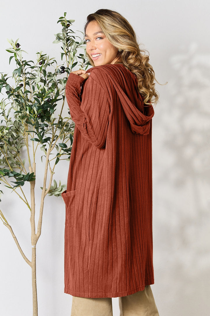 Wearing a warm smile, a person with long hair is dressed in the Basic Bae Full Size Hooded Sweater Cardigan, characterized by its opaque stretch fabric and rust color. They are standing beside a green plant against a light background.