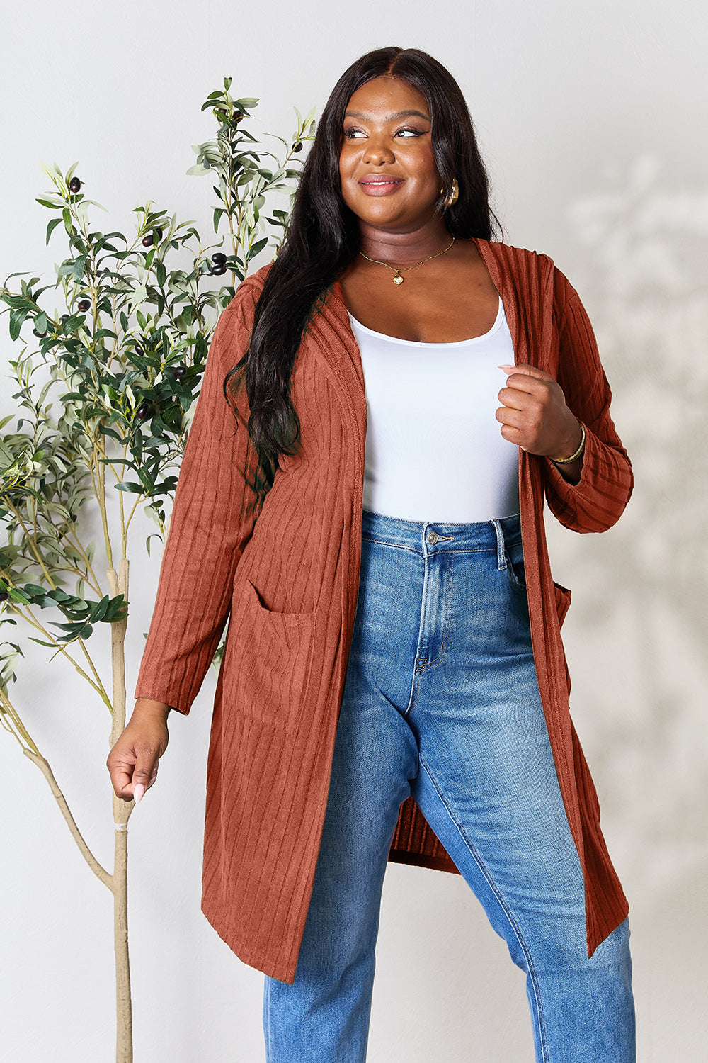 A woman stands smiling, wearing the Basic Bae Full Size Hooded Sweater Cardigan in a rust color over a white top and blue jeans. The cardigan's opaque stretch fabric fits comfortably as she poses with one hand on her hip in front of a leafy plant against a neutral background.