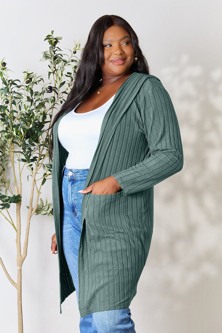 A person wearing the Basic Bae Full Size Hooded Sweater Cardigan in green, layered over a white top and blue jeans with an opaque stretch design, stands by an indoor plant.