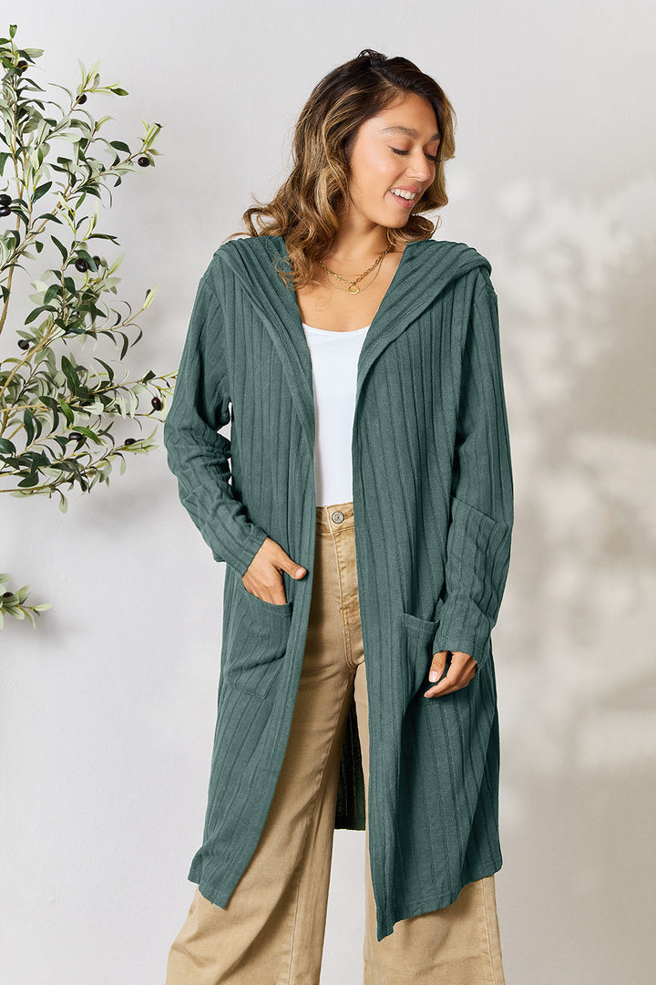 A person wearing the Basic Bae Full Size Hooded Sweater Cardigan in a long, green, opaque stretch design drapes it over a white top and beige pants. They stand near a leafy plant, smiling slightly while looking down. The cardigan features pockets and a hood, crafted for both comfort and style.