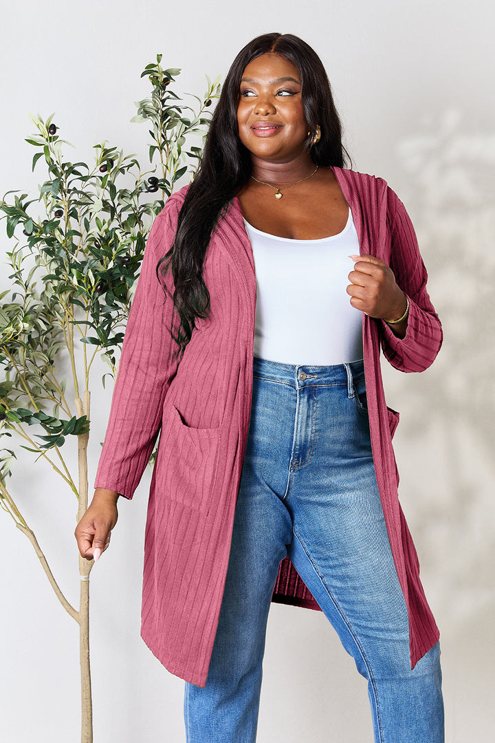 A person wearing a Basic Bae Full Size Hooded Sweater Cardigan in pink, along with a white top and blue jeans with opaque stretch, stands smiling near a plant.