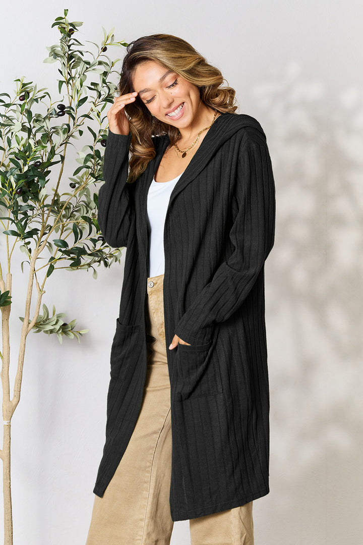 A woman stands smiling in a Basic Bae Full Size Hooded Sweater Cardigan, its long black design featuring pocketed sheer details, draped elegantly over a white top and beige pants. Her wavy hair cascades as she touches her head, with a potted plant in the background adding a touch of nature to the scene.