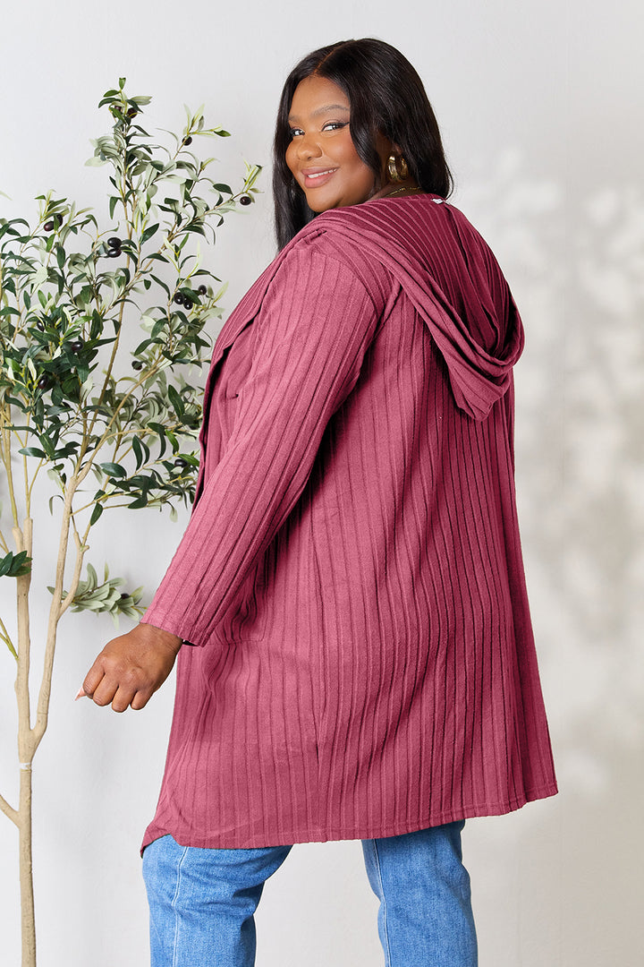 A person with long hair smiles while wearing the Basic Bae Full Size Hooded Sweater Cardigan, which is a cozy, opaque stretch maroon piece. They are looking over their shoulder and standing next to a small potted plant against a light-colored background.