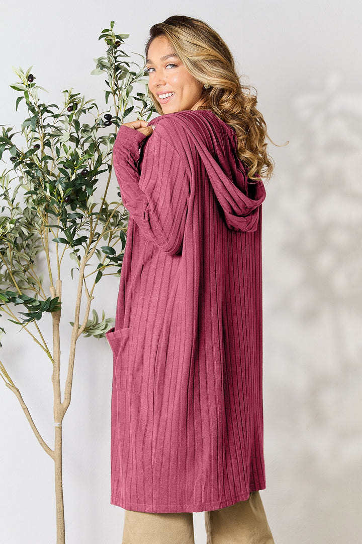 A person with long, wavy hair is smiling while looking over their shoulder, wearing the Basic Bae Full Size Hooded Sweater Cardigan—a long, ribbed maroon cardigan with a hood. This cozy piece is ideal for chilly days and can be machine washed in cold water. They're standing next to a small potted plant.