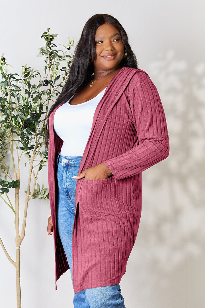 In a well-lit room, a woman wears the Basic Bae Full Size Hooded Sweater Cardigan in maroon, styled over a white top and blue jeans. She smiles while gazing to the side, with a small plant in the background providing a hint of nature to the setting.