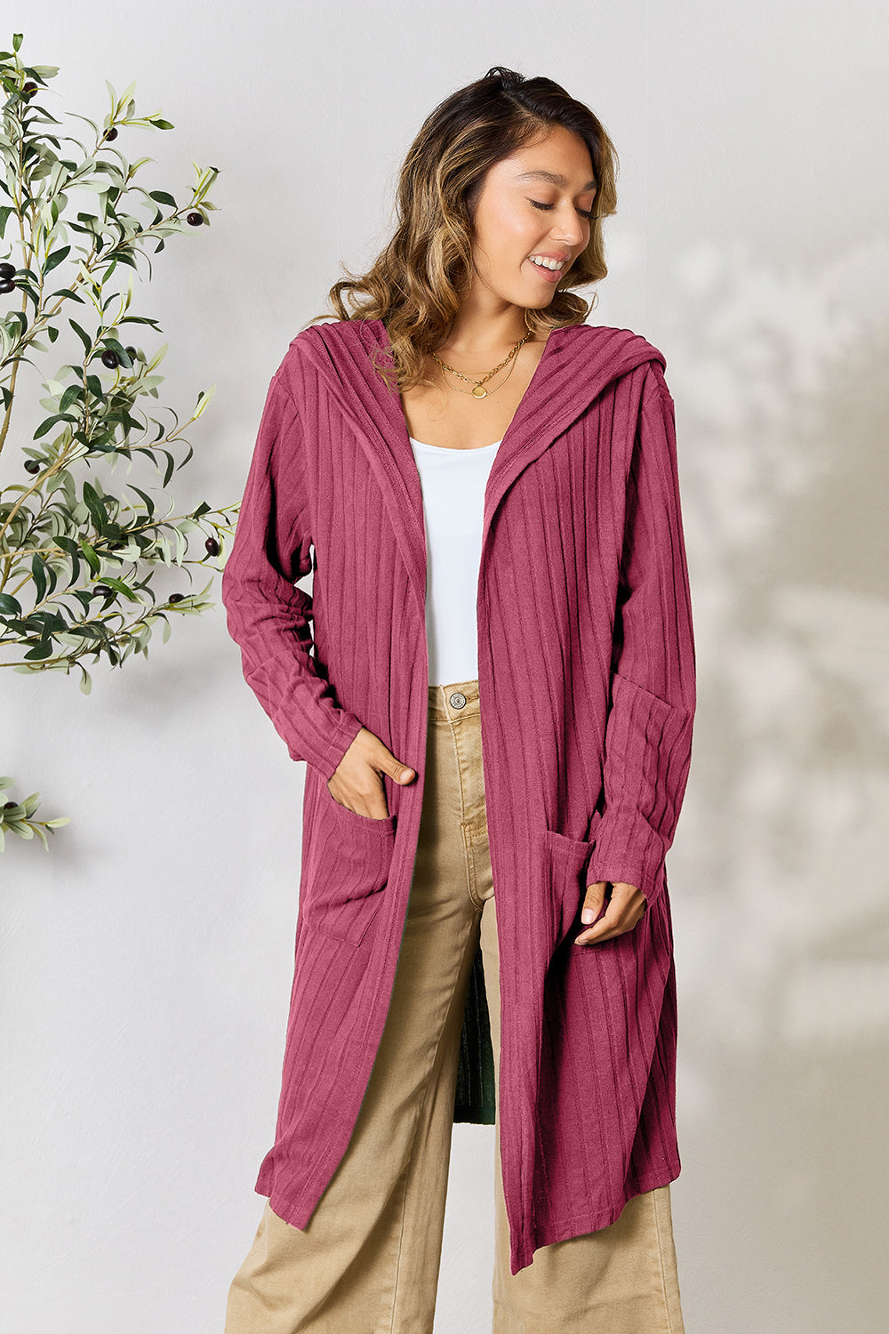 A person stands smiling, wearing the Basic Bae Full Size Hooded Sweater Cardigan in a maroon shade with sheer pocketed fabric. Underneath, they have on a white top and tan pants. In the background, a plant with green leaves enhances the cozy outfit that's perfect for any day out.