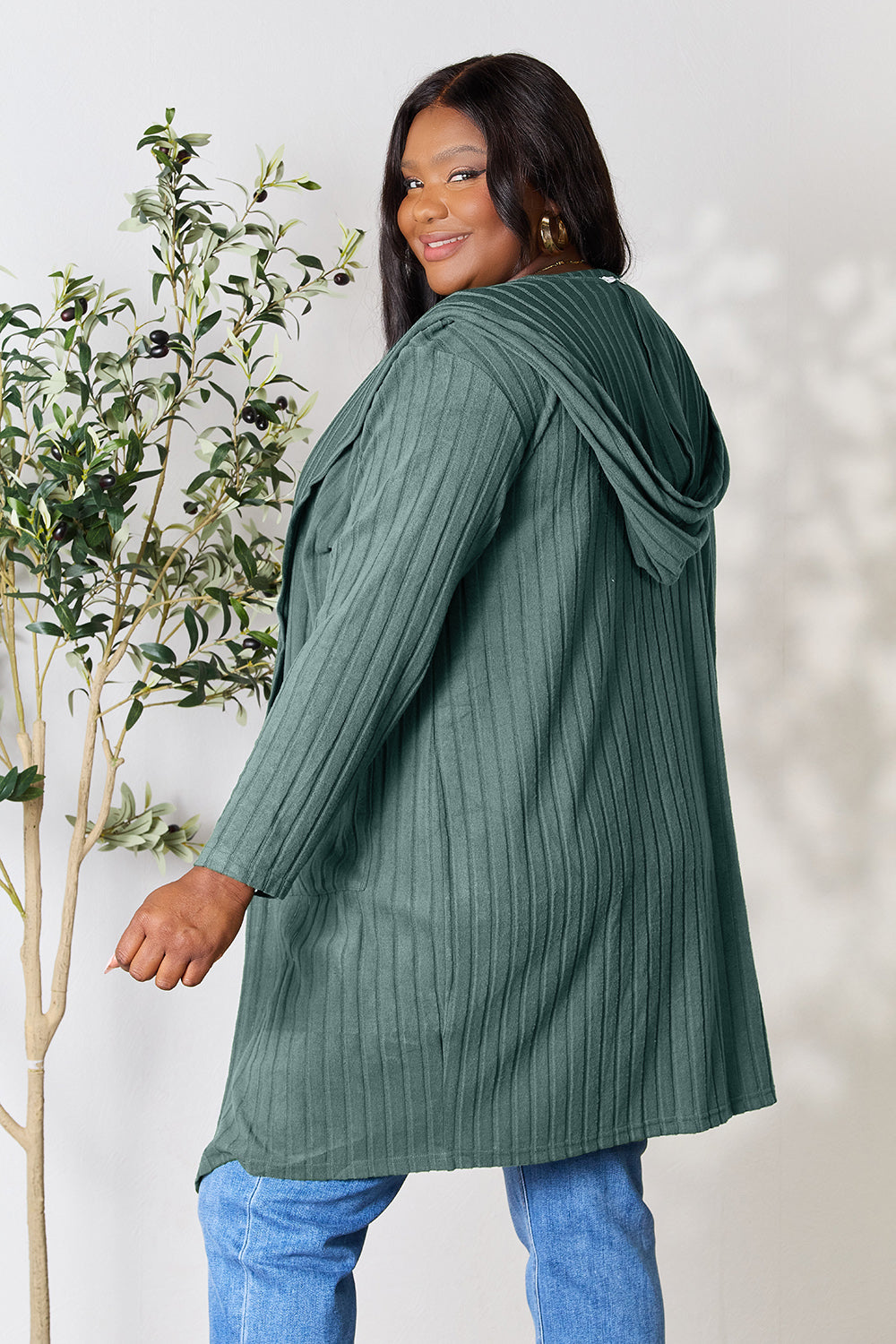 A person wearing the Basic Bae Full Size Hooded Sweater Cardigan in a long, dark green style, paired with blue jeans, poses confidently, smiling over their shoulder. A small leafy plant sits beside them against a plain light background.