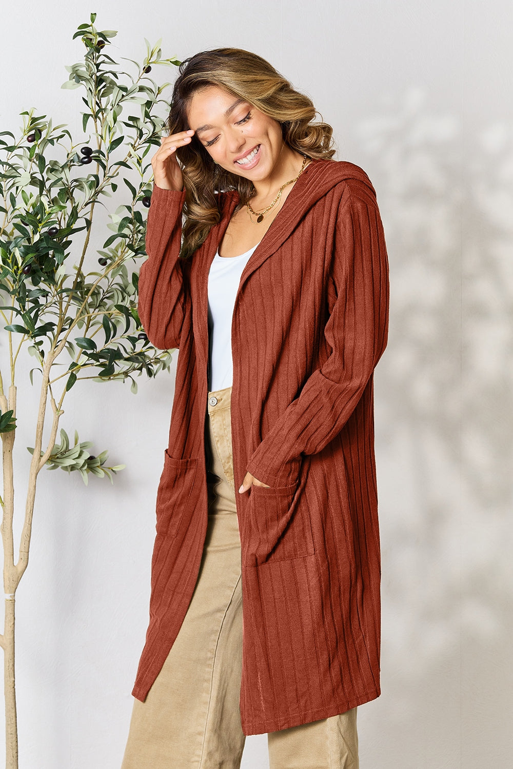 A woman stands smiling in the Basic Bae Full Size Hooded Sweater Cardigan, a cozy rust-colored sweater featuring convenient pockets. Crafted from comfortable opaque stretch fabric, it pairs perfectly with her white top and beige pants. A small indoor plant adds a touch of greenery to the background.