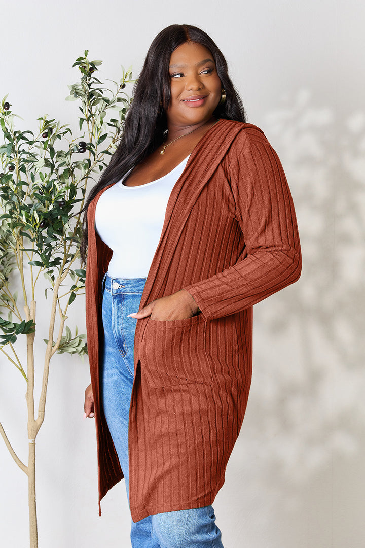 A person is smiling while wearing the Basic Bae Full Size Hooded Sweater Cardigan, a long, rust-colored layer over a white top and jeans. Standing in front of a light-colored wall with a small potted tree on the side, they enjoy the easy-care comfort of this opaque stretch fit cardigan—simply machine wash cold for convenience.