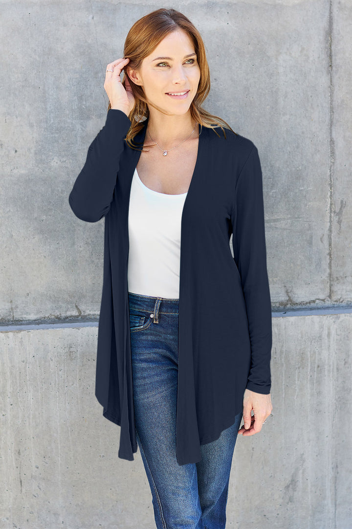 A woman with light brown hair stands against a concrete wall, wearing the Basic Bae Full Size Open Front Long Sleeve Cardigan in navy blue over a white top and blue jeans. Her outfit features a basic style crafted from slightly stretchy rayon spandex fabric. Smiling, she touches her hair with one hand, exuding effortless charm.