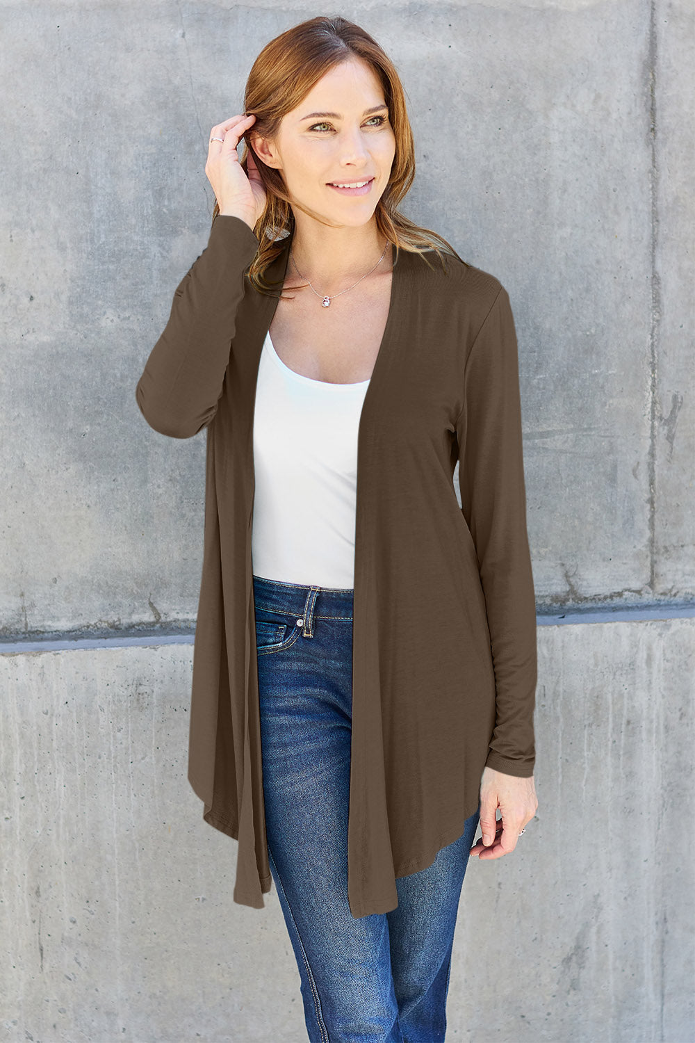 A woman with brown hair wearing a slightly stretchy white top, the Basic Bae Full Size Open Front Long Sleeve Cardigan in brown, and blue jeans crafted from rayon spandex stands in front of a gray concrete wall, smiling and touching her hair in a basic style.