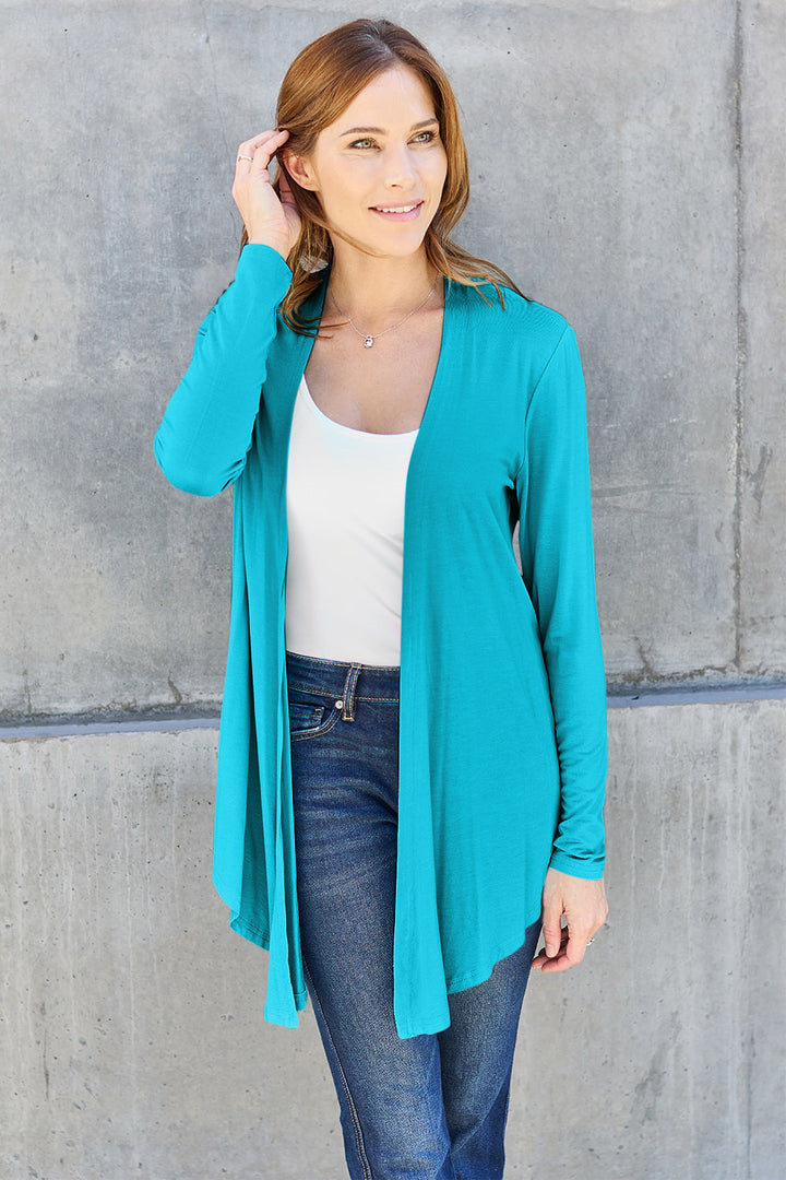 A woman with long hair, wearing the Basic Bae Full Size Open Front Long Sleeve Cardigan in turquoise—crafted from a slightly stretchy rayon spandex blend—over a white top and jeans poses against a gray concrete wall, touching her hair and smiling.