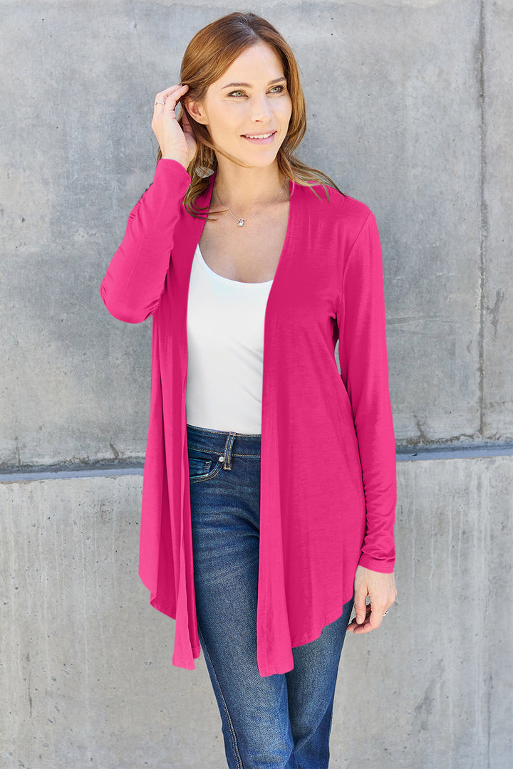 A woman stands against a gray concrete wall, wearing the Basic Bae Full Size Open Front Long Sleeve Cardigan in bright pink rayon spandex. She pairs it with a white top and blue jeans. Her light brown hair frames her gently smiling face as she lightly touches her hair, showcasing her effortlessly basic style.