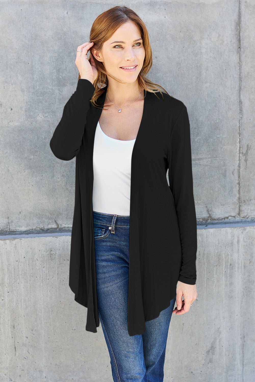 A woman stands in front of a concrete wall, showcasing a classic look with the Basic Bae Full Size Open Front Long Sleeve Cardigan in black over a white top and blue jeans. She casually touches her hair and smiles, exuding a relaxed and confident posture.