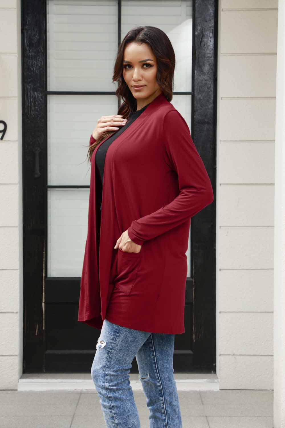 A confident woman wearing the Basic Bae Full Size Open Front Long Sleeve Cardigan with Pockets in red pairs it with blue jeans. Crafted from imported polyester for easy machine washing, she stands with one hand in her pocket, posing in front of a black door and a light-colored wall while gazing at the camera.