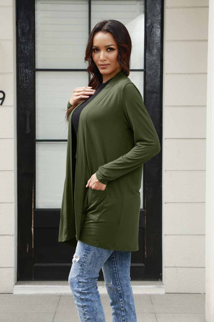 A woman stands confidently in front of a black door, her long hair flowing as she wears the Basic Bae Full Size Open Front Long Sleeve Cardigan with Pockets in green, paired with a black top and ripped jeans. One hand rests on the polyester cardigan while the other is casually tucked into her pocket.