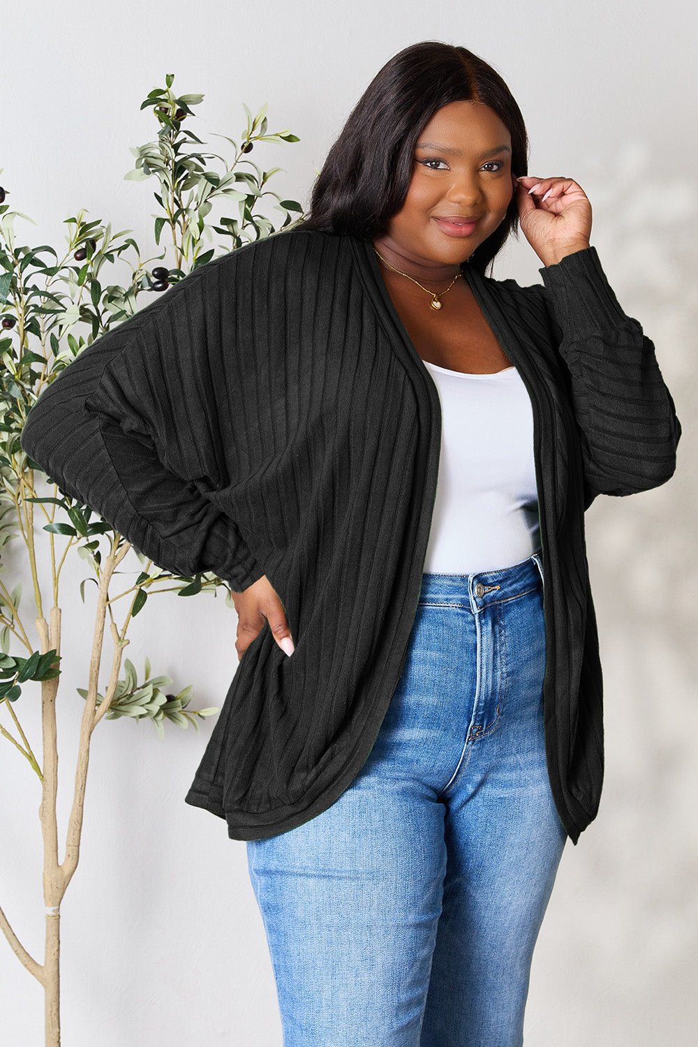 A woman stands against a light background, sporting the Basic Bae Full Size Ribbed Cocoon Cardigan in black over a white top and blue jeans. With her hand gently touching her hair and a leafy plant beside her, she smiles softly, perfectly capturing the essence of basic style.