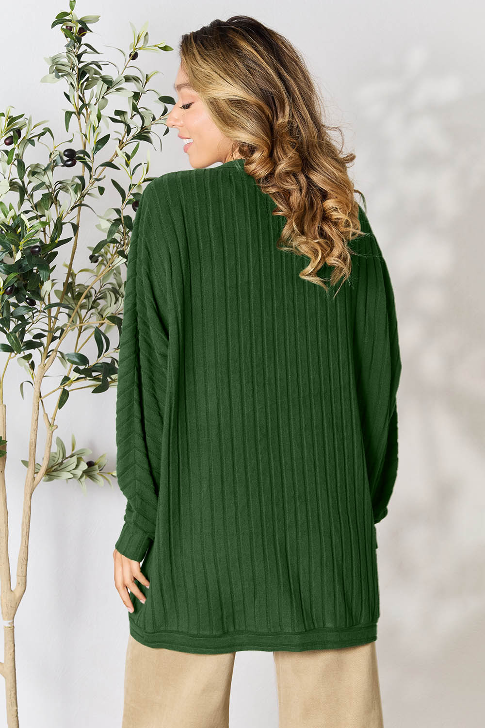 A woman with long wavy hair is turned away, wearing the oversized Basic Bae Full Size Ribbed Cocoon Cardigan made from slightly stretchy polyester. She stands next to a potted plant with green leaves, against a neutral background.