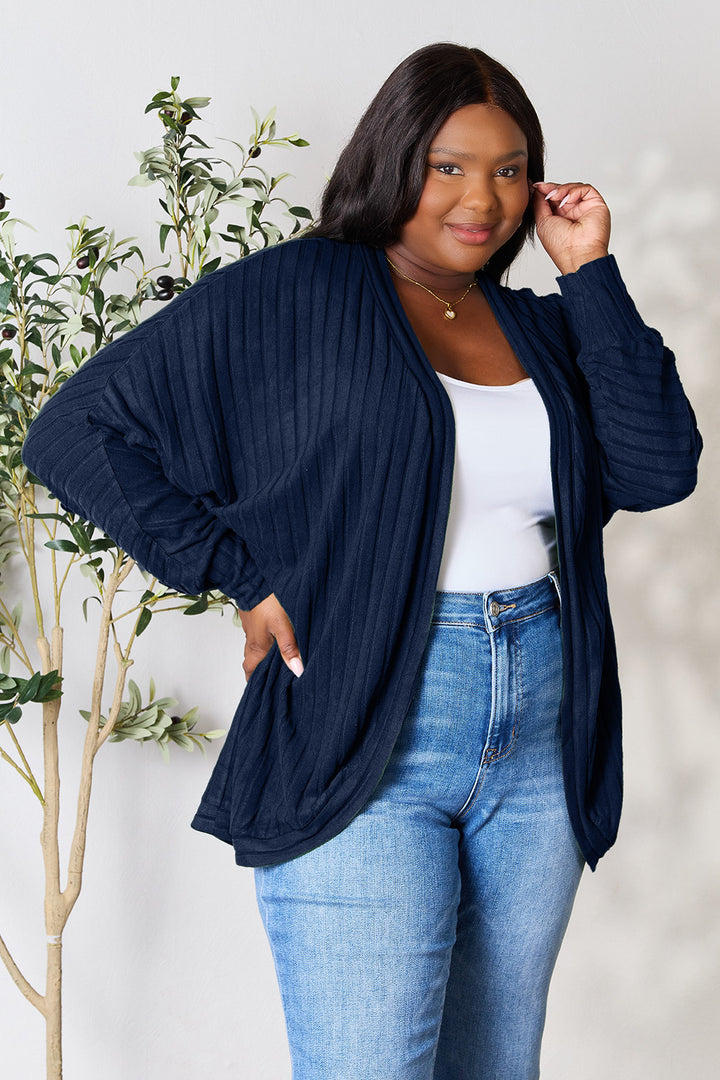 A person stands confidently, posing with one hand on their hip and the other touching their hair. They wear the Basic Bae Full Size Ribbed Cocoon Cardigan in navy blue over a basic-style white top and blue jeans. A plant is visible in the background.