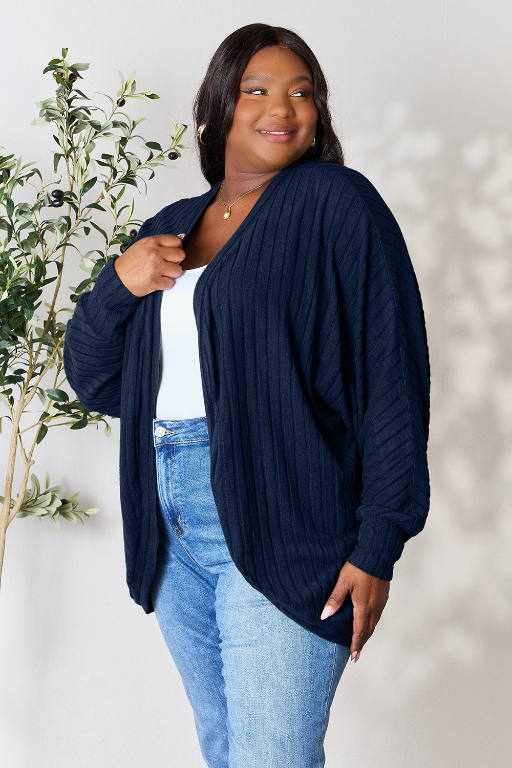 Wearing the Basic Bae Full Size Ribbed Cocoon Cardigan in a slightly stretchy navy blue polyester, a person poses confidently in front of a light background with jeans and a small plant by their side. The cardigan's ribbed texture complements their basic style chicness as they smile gently while looking to the side.