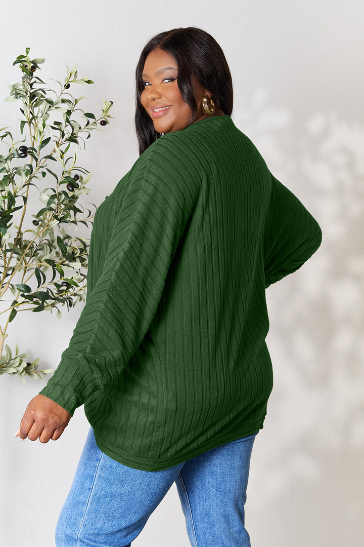 A person wearing a dark green Basic Bae Full Size Ribbed Cocoon Cardigan and blue jeans is smiling and looking over their shoulder. A small leafy plant adds a touch of nature to this basic style scene.