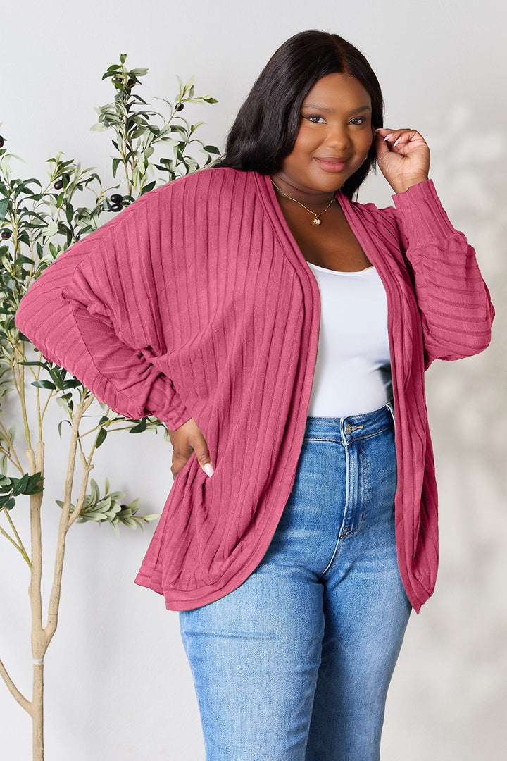 Dressed in the Basic Bae Full Size Ribbed Cocoon Cardigan, a person confidently stands against a light grey background adorned with a leafy plant. The pink cardigan, made from slightly stretchy polyester, is stylishly paired with a white top and blue jeans. Embracing a basic style, they smile warmly while touching their hair.