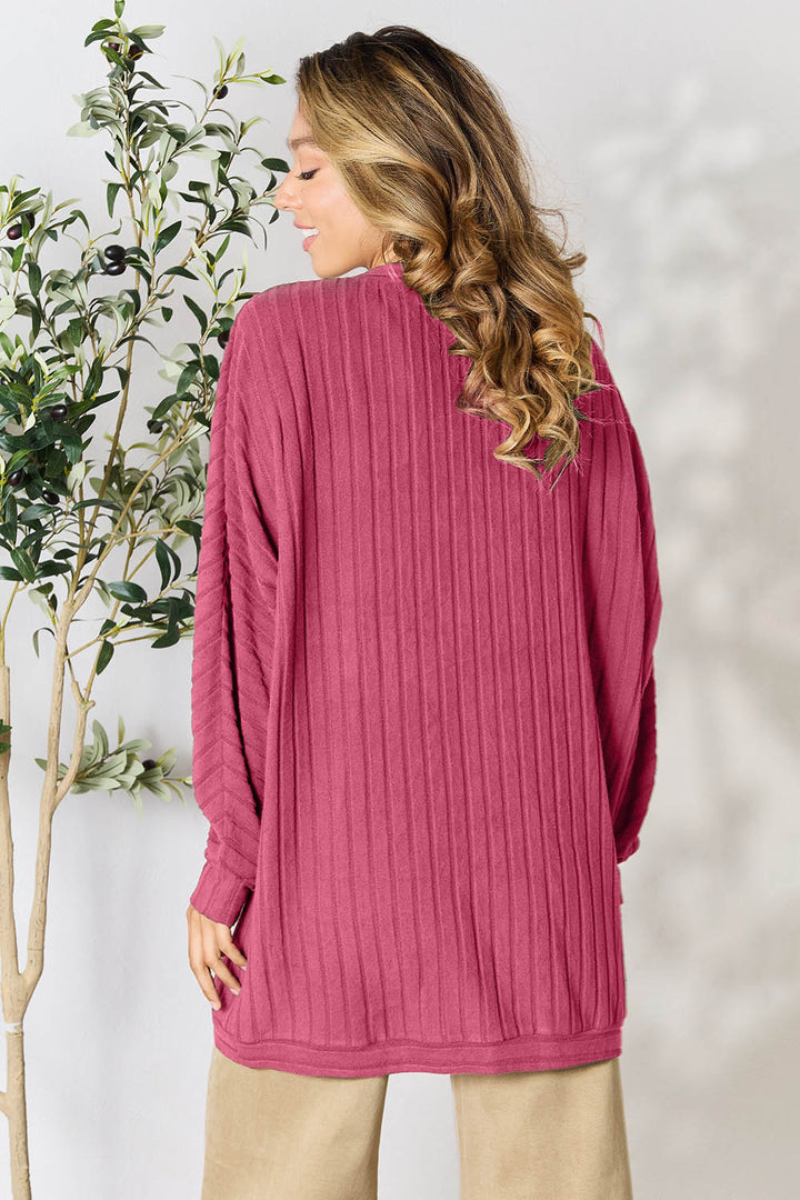 A person with long, wavy hair is standing sideways in the Basic Bae Full Size Ribbed Cocoon Cardigan, which is a loose, slightly stretchy pink sweater made from soft polyester. They are also wearing beige pants and embody a basic style next to a leafy plant against a light-colored background.