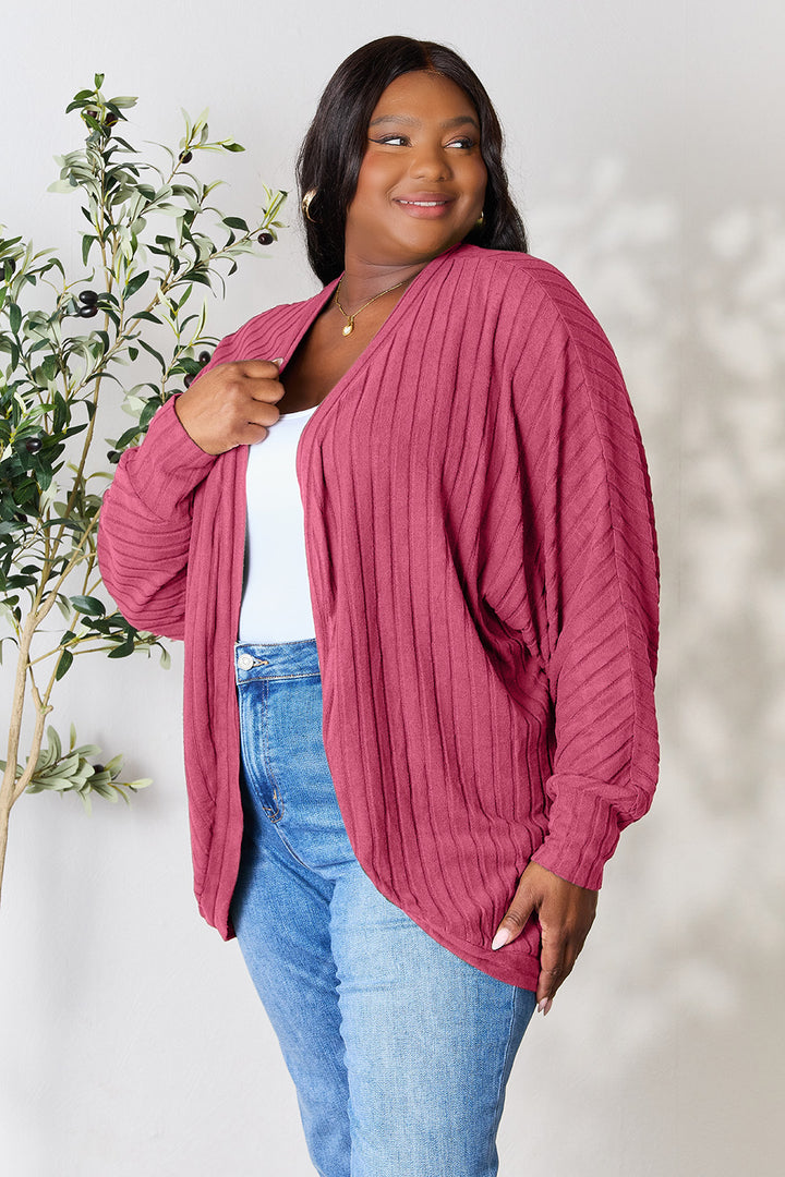 A person wearing the Basic Bae Full Size Ribbed Cocoon Cardigan, a long-sleeve, pink polyester piece with a textured and slightly stretchy design, over a white top and blue jeans smiles while standing near a leafy plant. The neutral background highlights the basic style of the outfit.